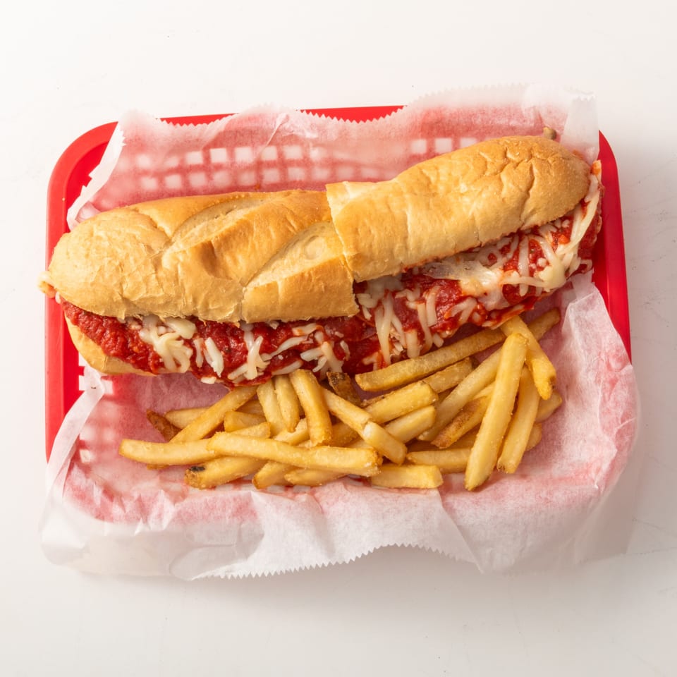Meatball sub with melted cheese and marinara sauce, served with golden french fries in a red basket.