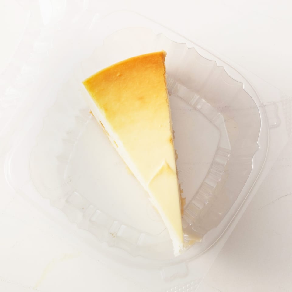 A slice of plain cheesecake with a golden-brown top sits in a clear plastic container.