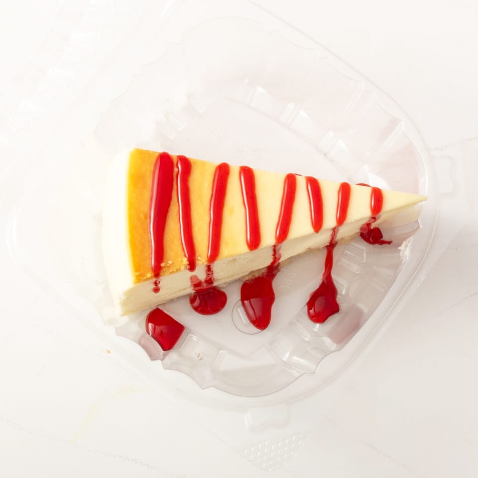 Slice of cheesecake with red raspberry drizzle in a clear plastic container.