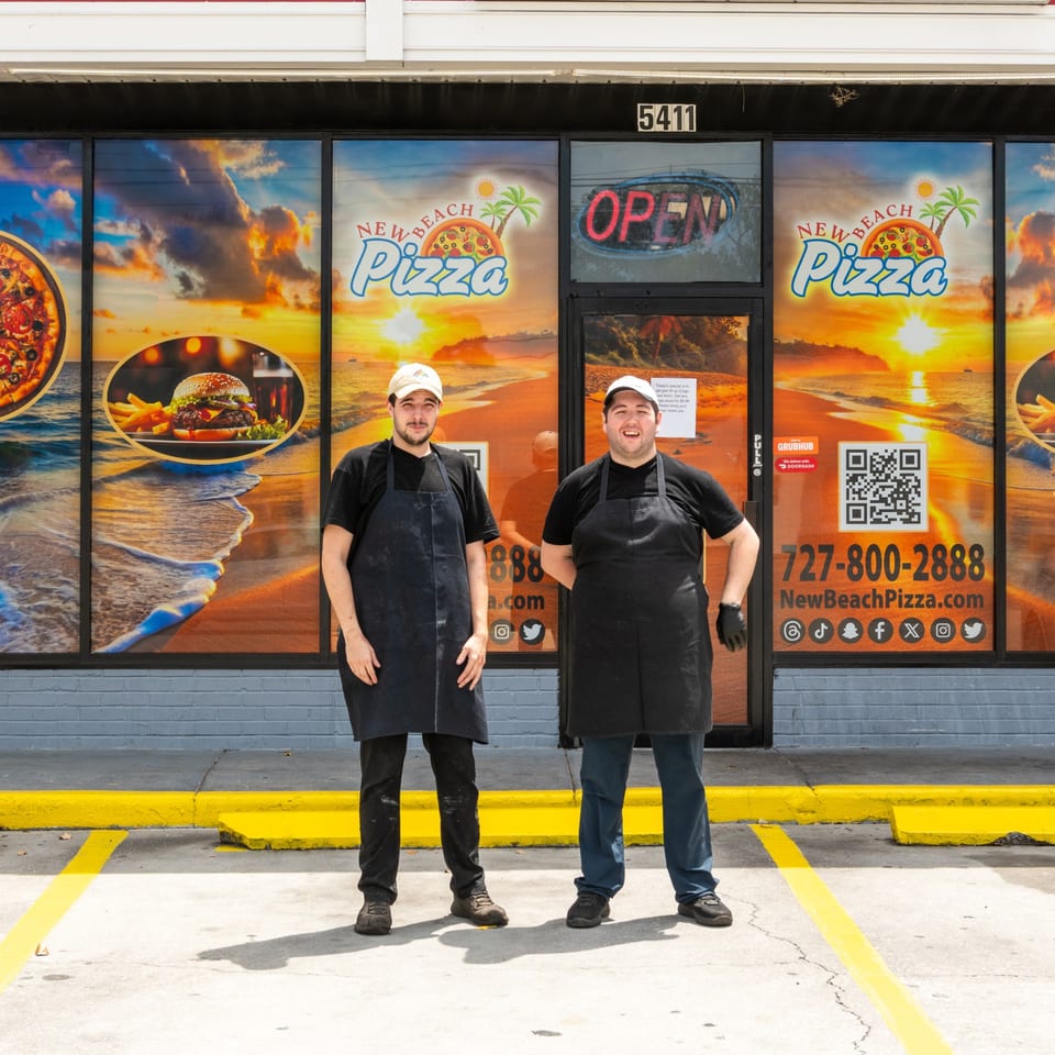 Two men in aprons stand outside New Beach Pizza, with sunset beach window decals.