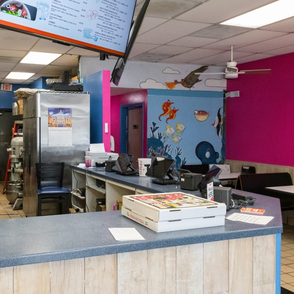 A restaurant counter with two pizza boxes, a menu screen, and colorful murals on the walls.